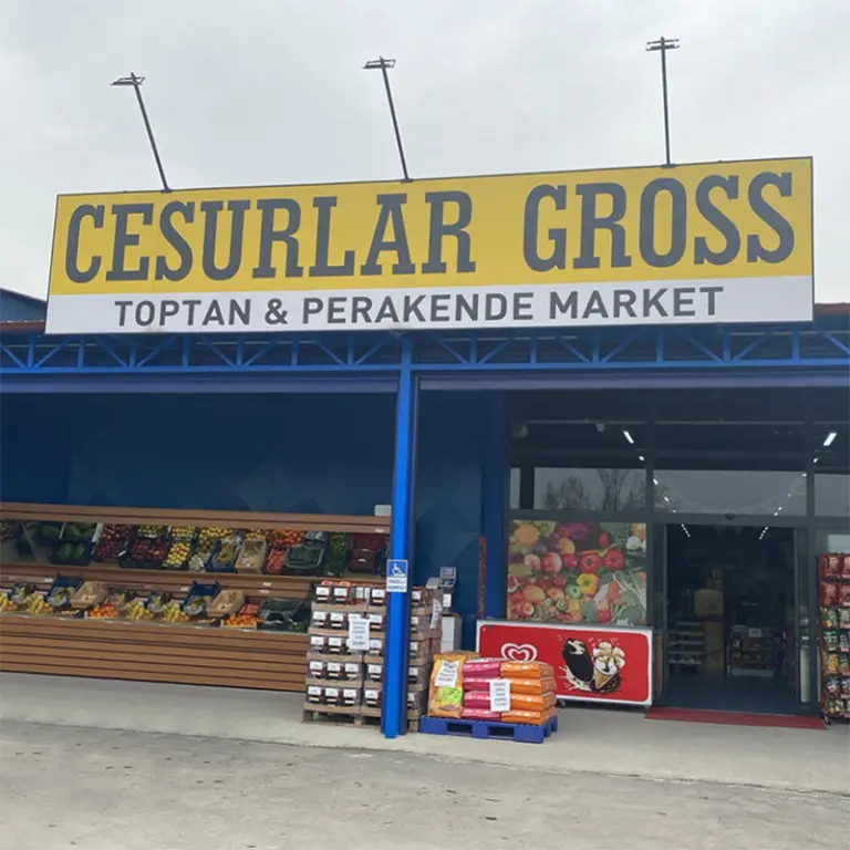 Cesurlar Gross mağaza önü: sarı tabela, mavi çelik saçak ve meyve-sebze tezgâhları